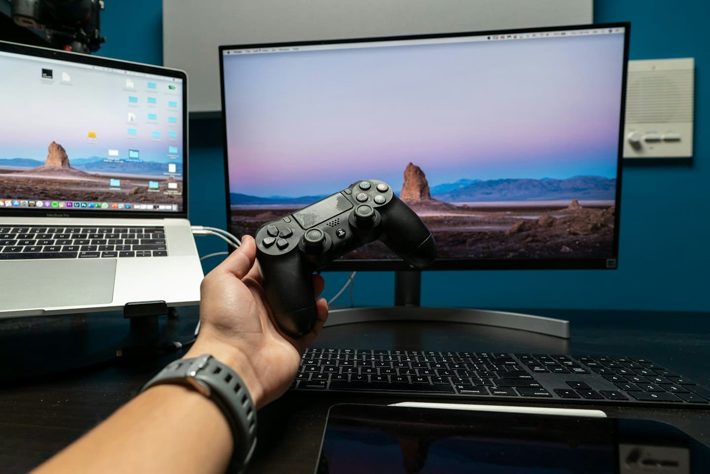 Mano che tiene un controller di videogiochi vicino a un laptop e un monitor.